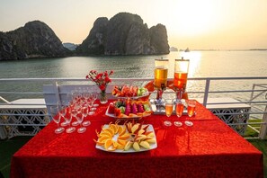 A table set with fruit, glasses, and a drink dispenser, overlooking a body of water with rock formations. - Halong Royal Palace Cruise (Tuan Chau)
