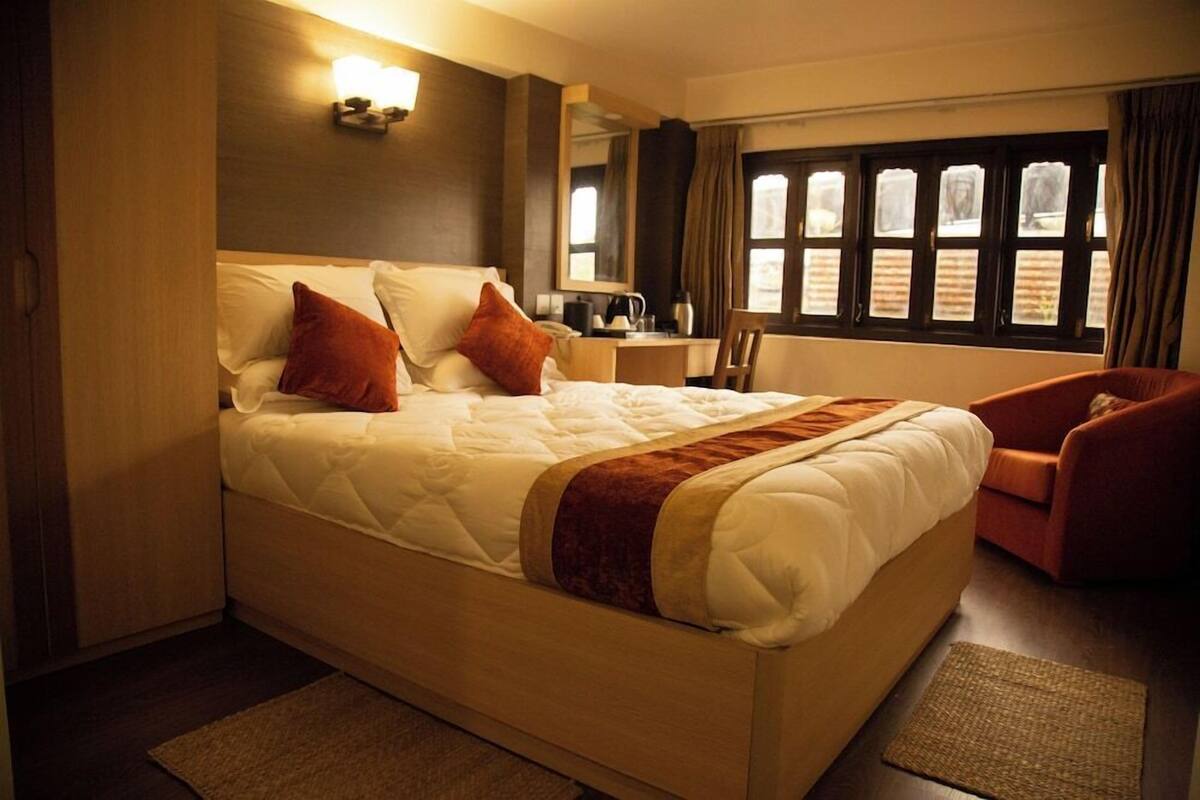 A hotel room with a large bed, a desk, a chair, a window with curtains, and a wall-mounted light fixture.