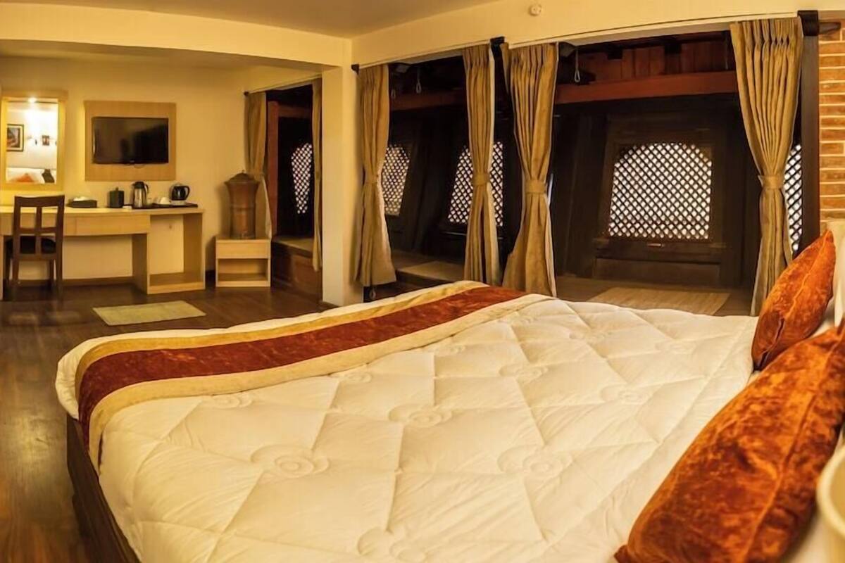 A bedroom with a large bed, a desk, and a chair. There are curtains and a window with a lattice design.