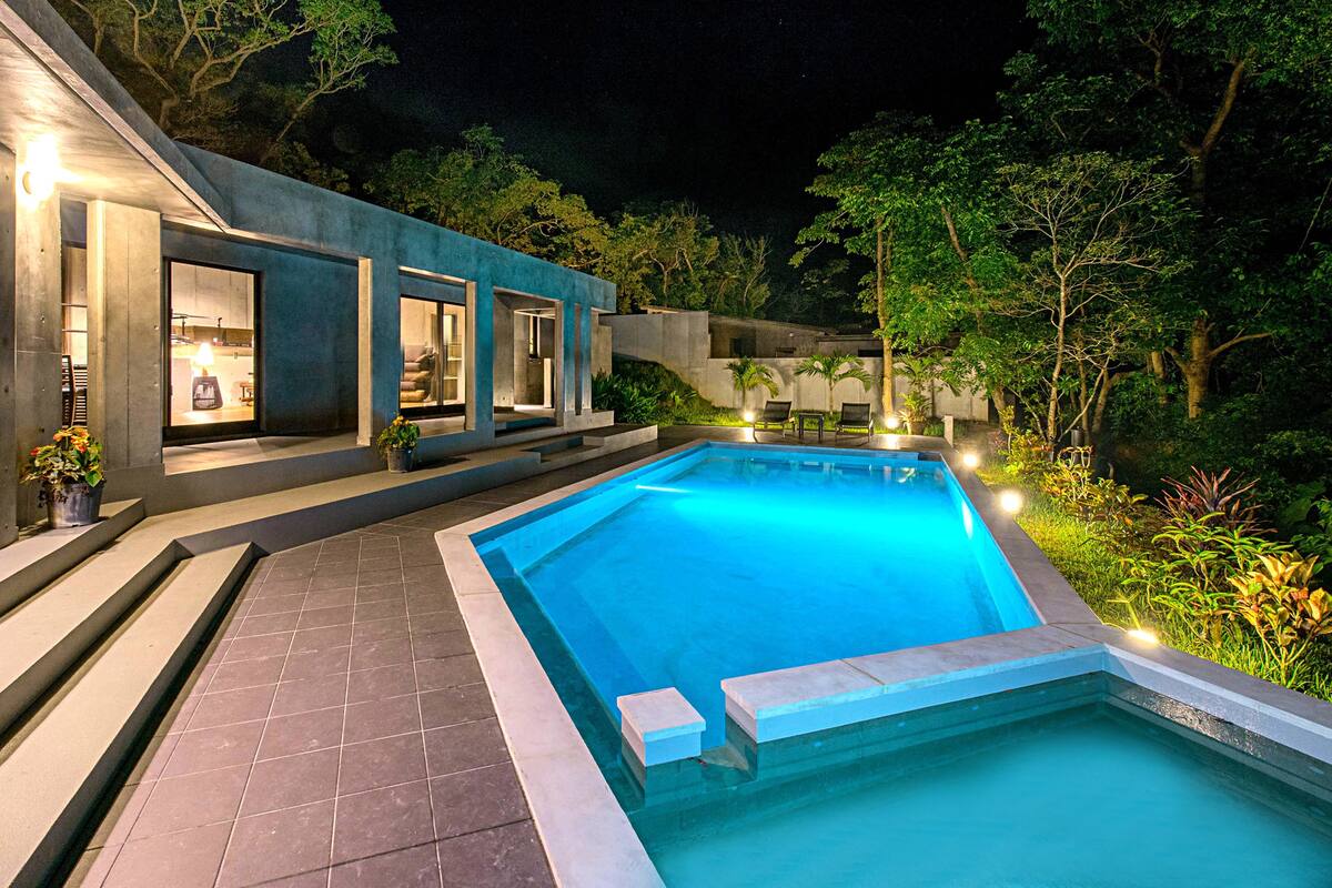 A modern outdoor pool area with a clear blue pool, surrounded by a well-lit patio with steps leading to a covered seating area.