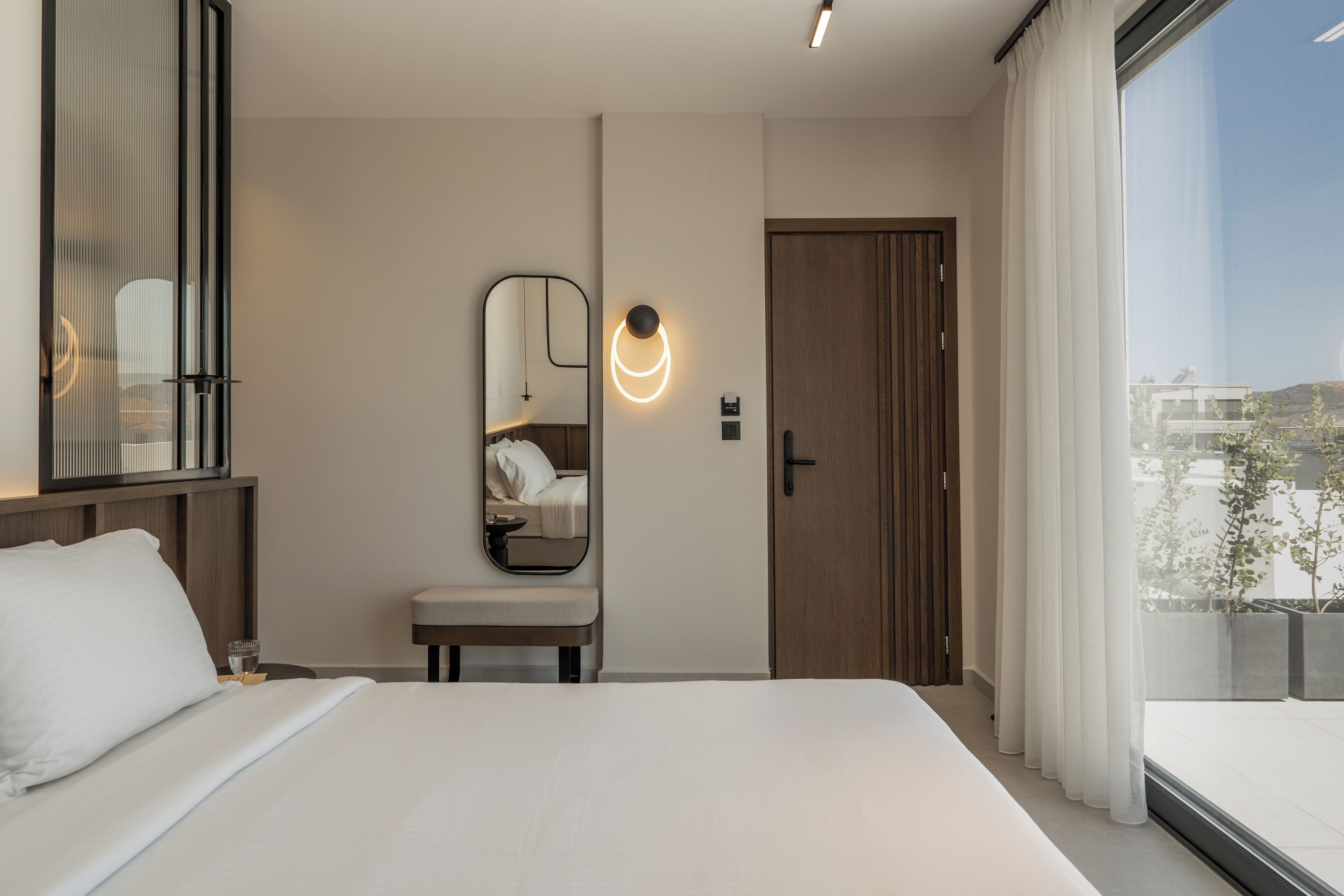 A modern hotel room with a large bed, a bedside table, a mirror, a door, and a window with curtains.