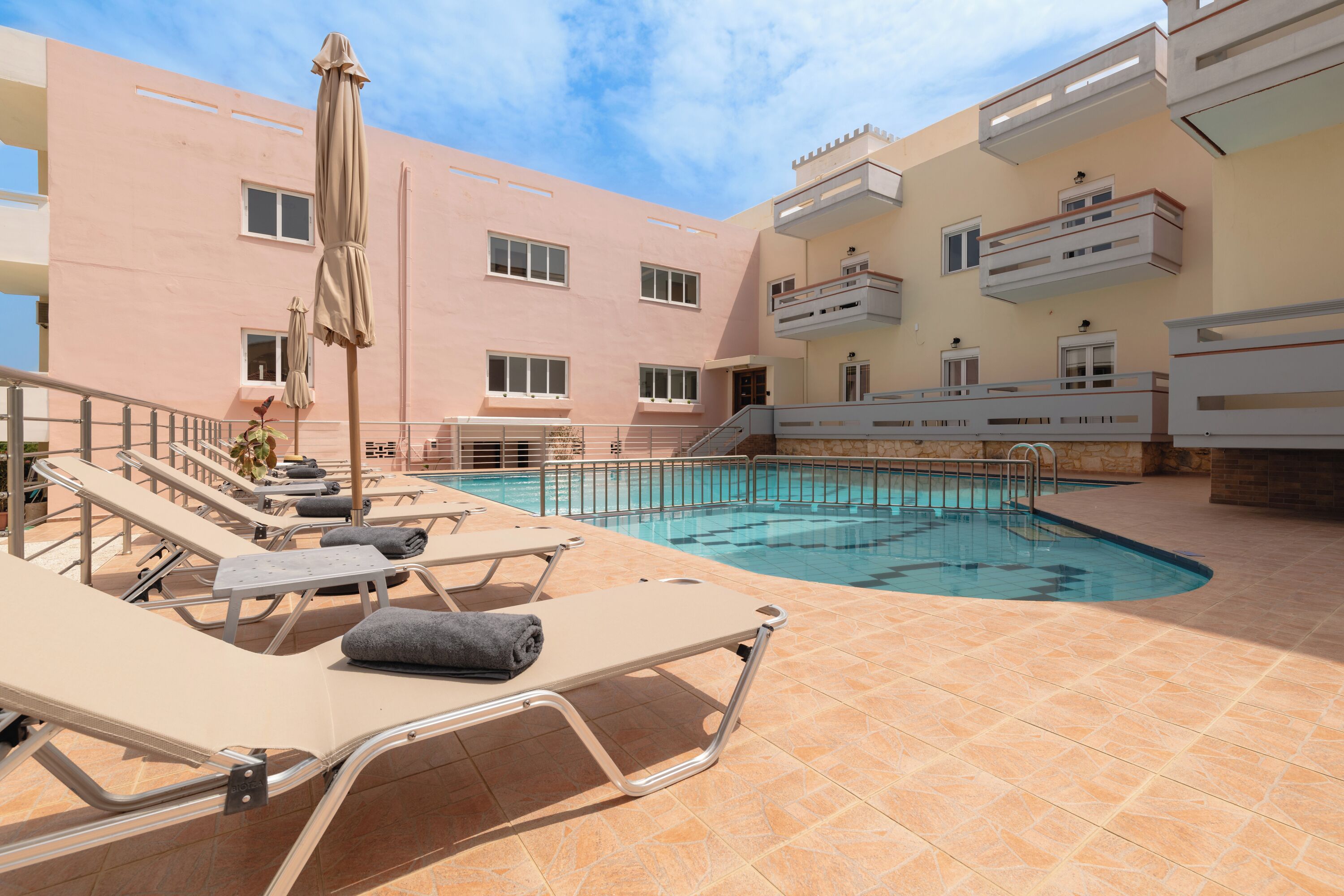 A pool area with lounge chairs and umbrellas, a building with balconies, and a clear sky.
