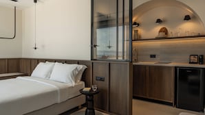Quarto de hotel moderno com cama, mesa de cabeceira e área de kitchenette com frigorífico e bancada.