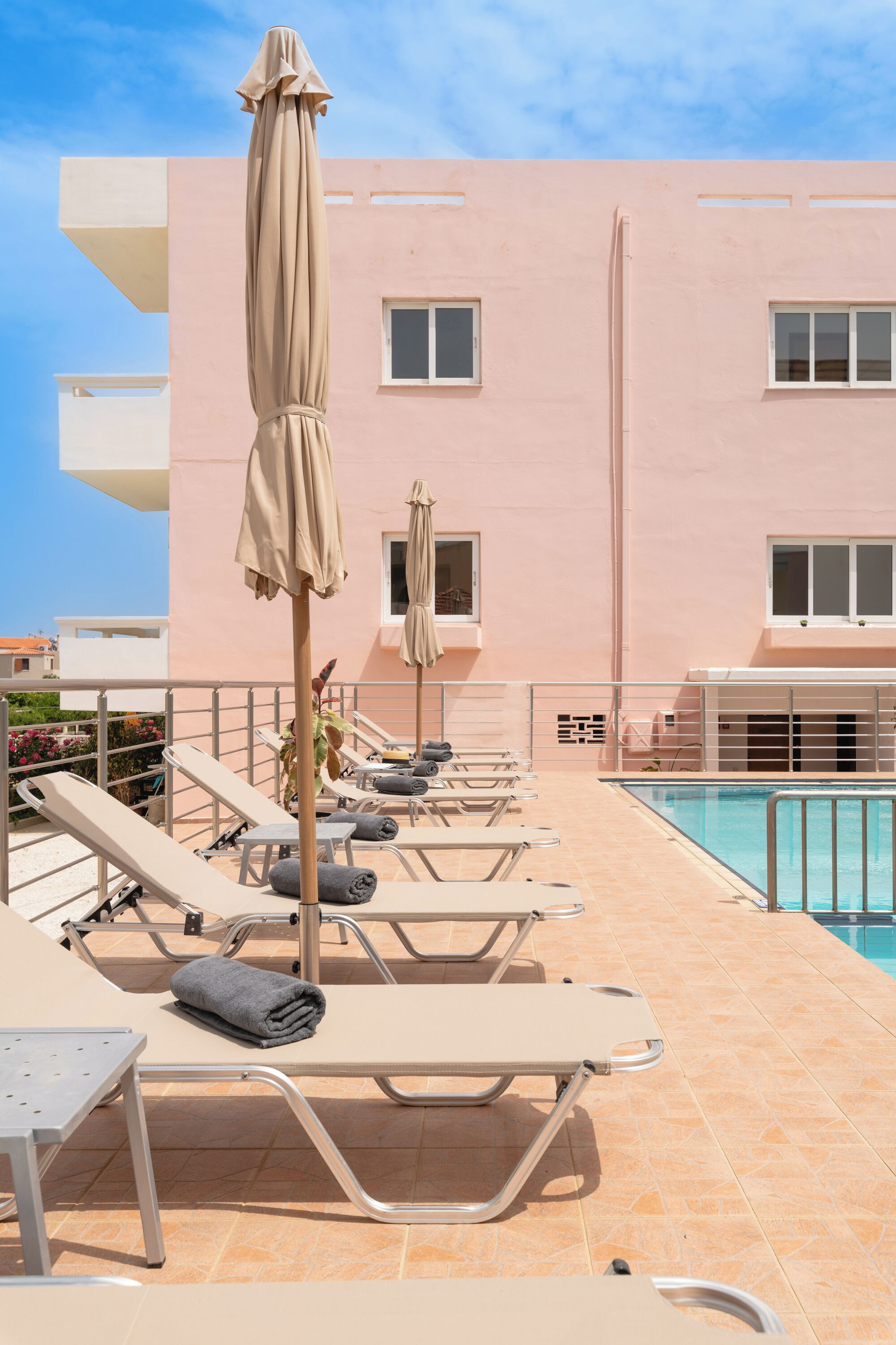 Uma área à beira da piscina com espreguiçadeiras e guarda-sóis, uma piscina e um edifício rosa ao fundo.