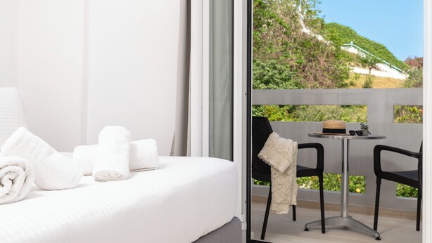 Quarto de hotel com cama, toalhas brancas, uma mesinha com chapéu e óculos de sol, e vista para uma encosta verde.