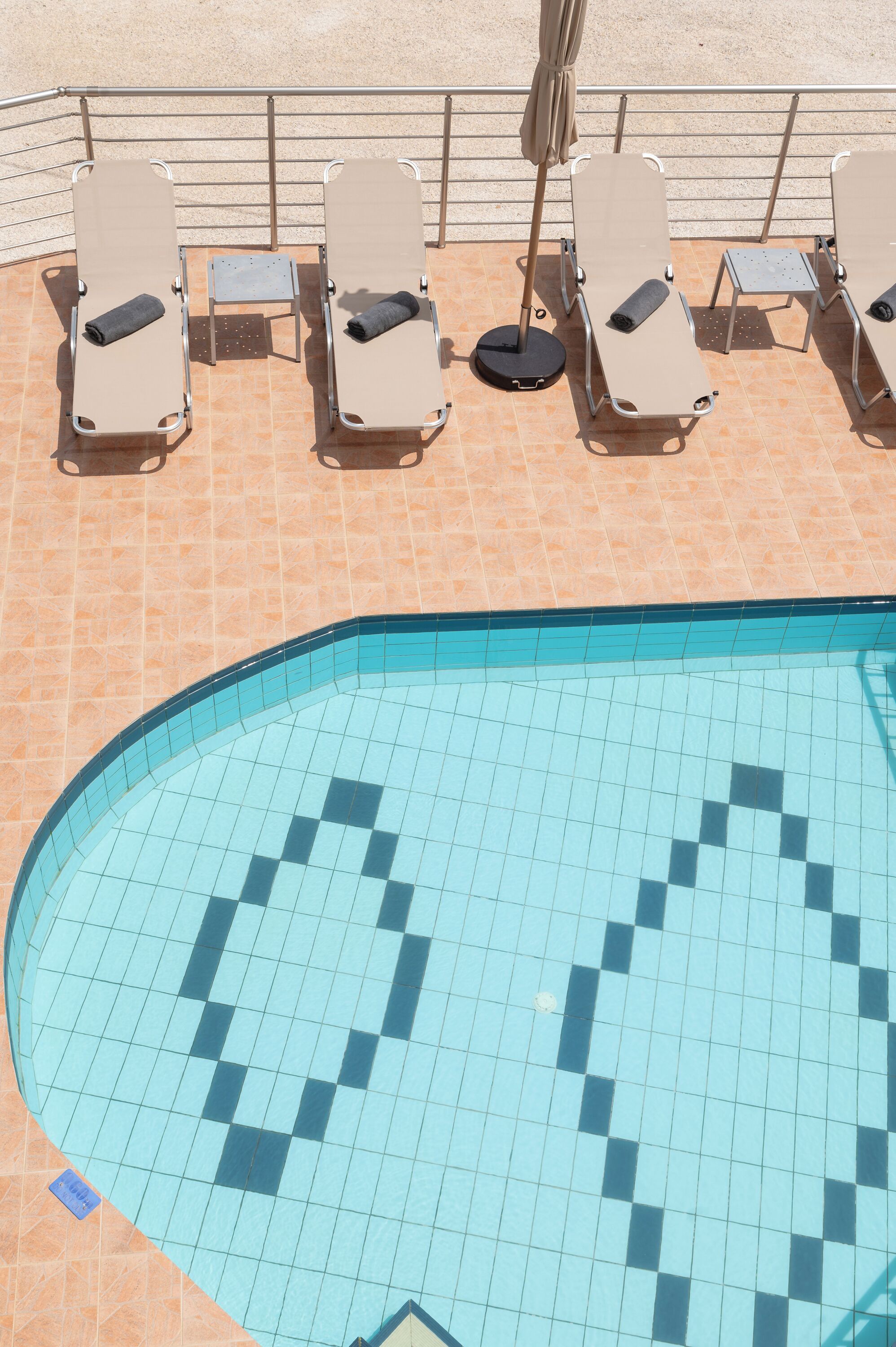 Uma área à beira da piscina com espreguiçadeiras e uma pequena mesa, cercada por um deck revestido de azulejos.