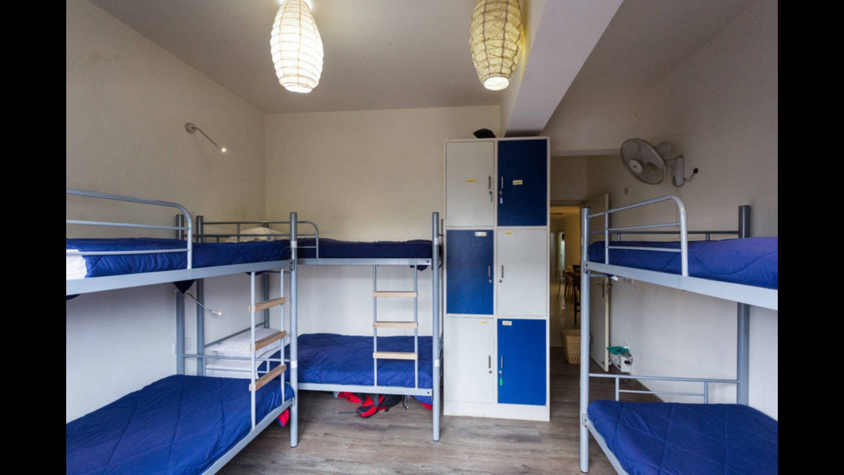 A dormitory room with bunk beds, a wardrobe, and a fan.