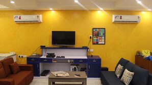 A living room with a TV, blue and brown furniture, and two air conditioners on the wall. - Joey's Hostel (New Delhi)