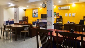 A modern dining area with a yellow accent wall, a clock, and a whiteboard with writing. - Joey's Hostel (New Delhi)