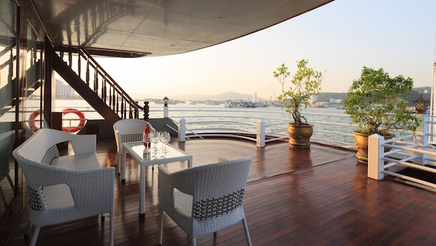 A deck with wicker chairs and a table, overlooking a body of water with boats and a distant cityscape. - Halong Silversea Cruise (Tuan Chau)