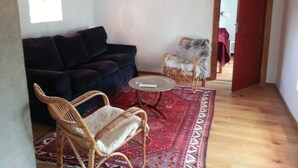 Een ruimtelijke zithoek met een donkerblauwe bank, een rieten fauteuil met een wit kussen, een klein rond tafeltje en een vloerkleed met een rood patroon.
