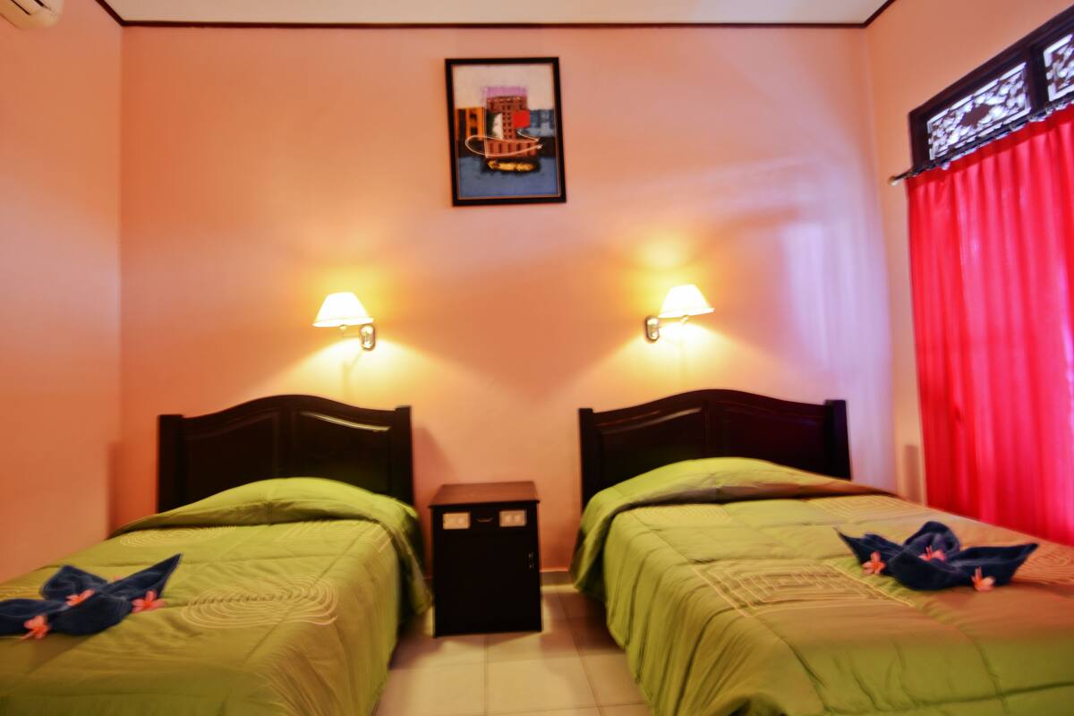 A room with two beds, each with green bedding and a headboard, a small nightstand between them, and a framed picture on the wall.