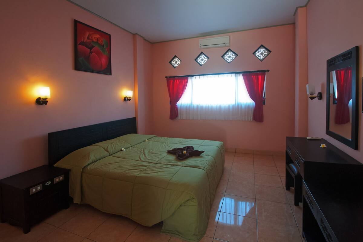 A bedroom with a bed, nightstands, a dresser, a window with red curtains, and a wall-mounted air conditioner.