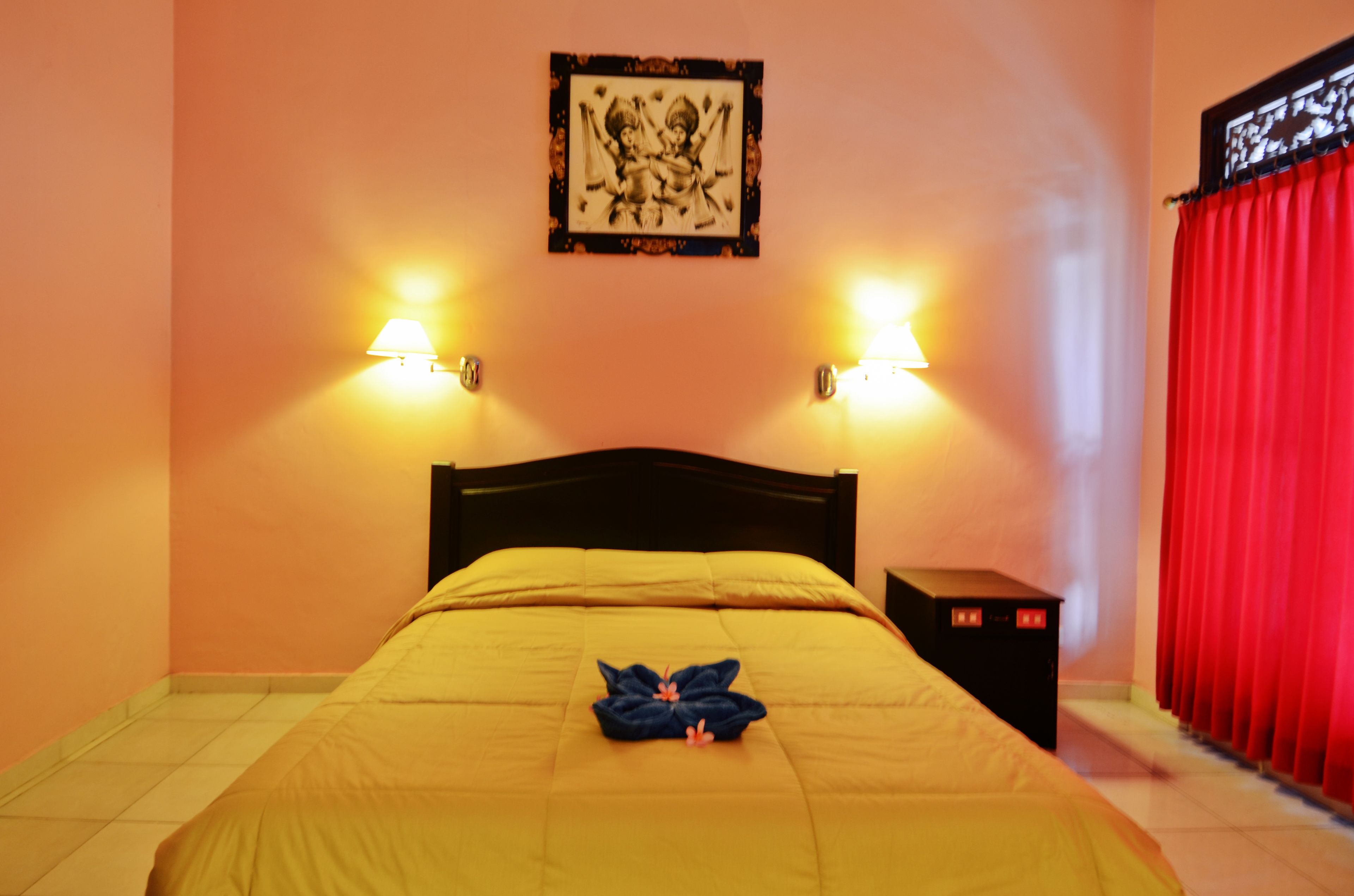 A hotel room with a bed, two wall-mounted lamps, a red curtain, and a framed picture on the wall.