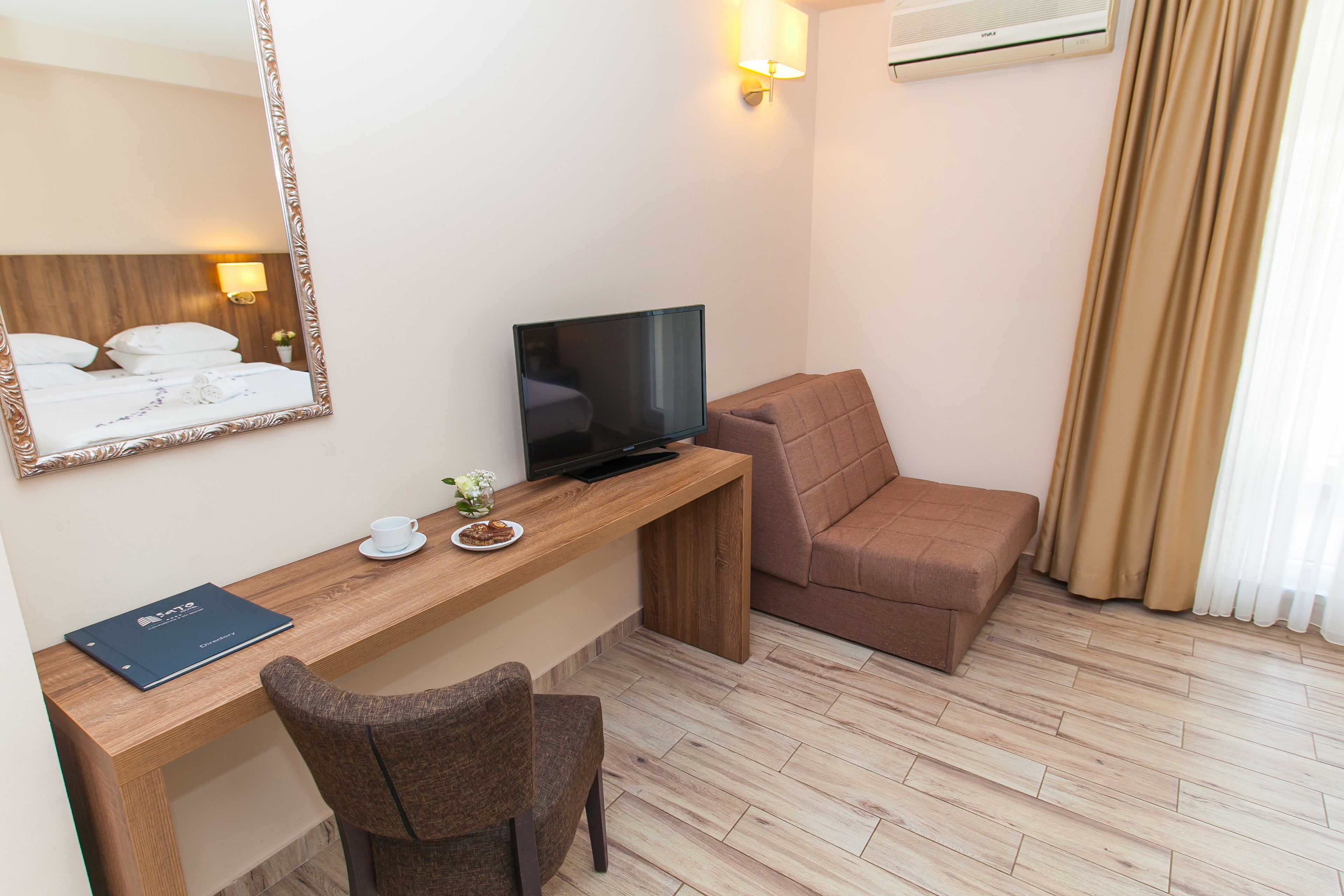 Ett hotellrum med ett skrivbord i trä, en tv, en brun soffa, en stol, ett litet bord med en kopp och en tallrik samt en spegel på väggen.