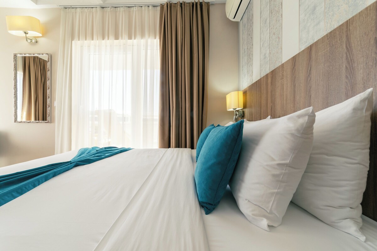A hotel room with a bed, curtains, a lamp, a mirror, and a wall-mounted air conditioner.