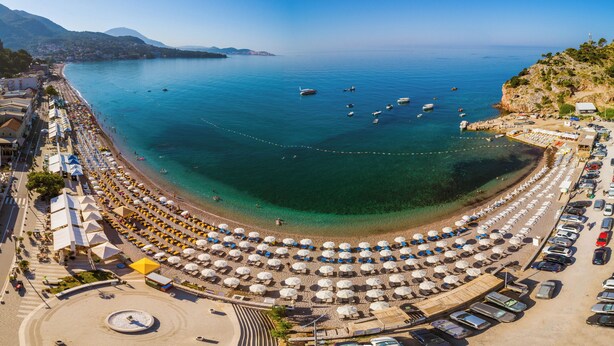A coastal beach with rows of sunbeds and umbrellas, a parking area, and boats in the water. - Hotel Sato (Sutomore)