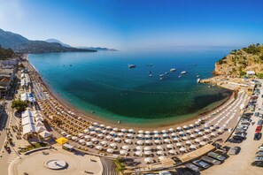A coastal beach with rows of sunbeds and umbrellas, a parking area, and boats in the water. - Hotel Sato (Sutomore)
