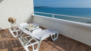 A balcony with two white sun loungers, a small table with a hat and a bottle, and a view of the sea and distant land. - Hotel Sato (Sutomore)