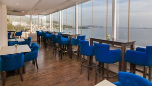 Un restaurant avec des chaises et des tables bleues, de grandes fenêtres donnant sur la mer et une ville située au loin.