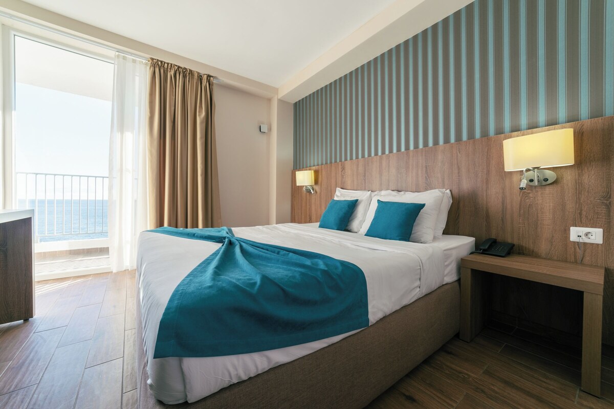 A hotel room with a bed, bedside table, lamp, and a view of the outdoors.