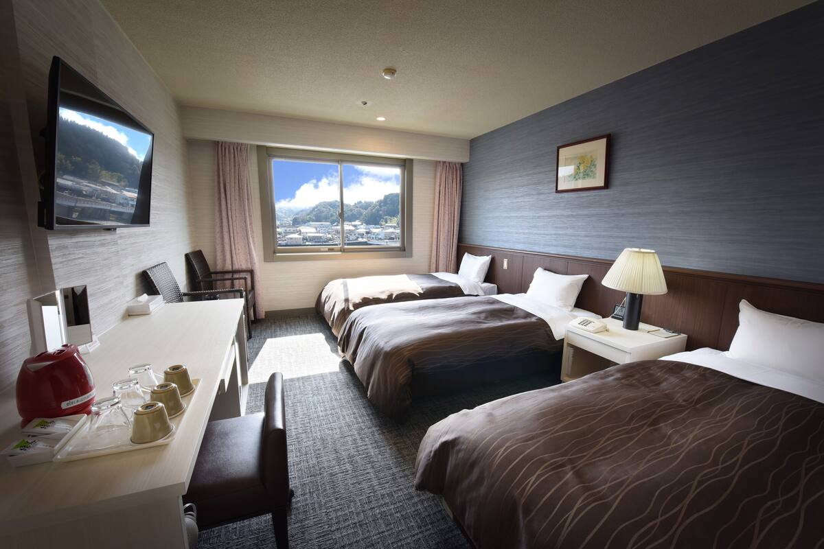 Een hotelkamer met twee bedden, een bureau met een rode waterkoker, een aan de muur bevestigde televisie en een groot raam met uitzicht op de stad.