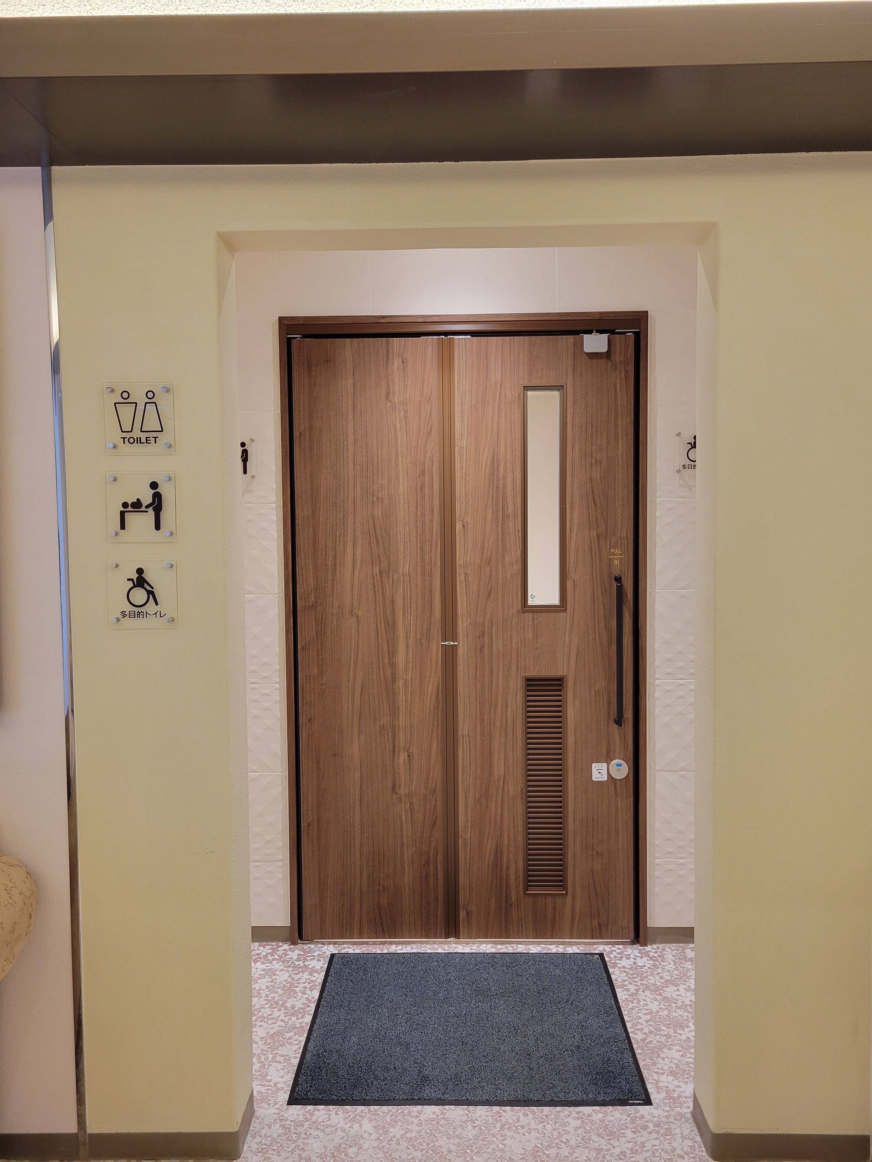 Puerta doble de madera con un espejo vertical, un felpudo y tres letreros de baño en la pared.