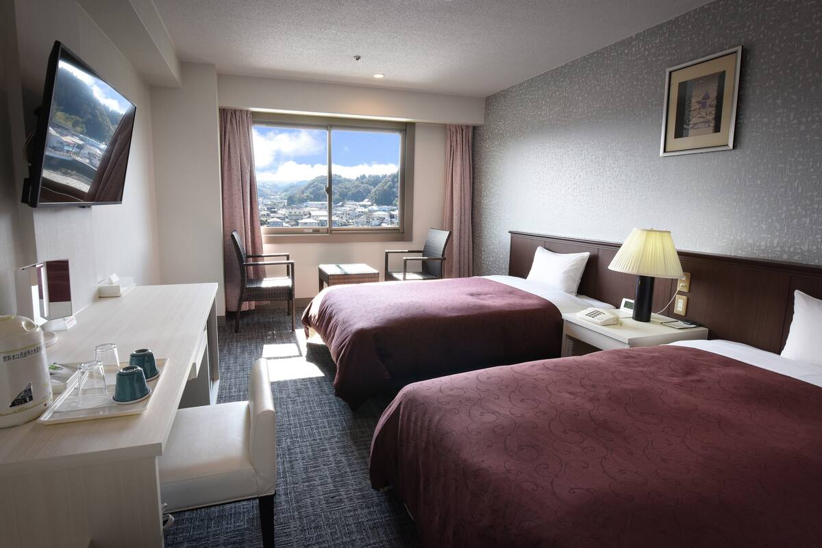 Een hotelkamer met twee bedden, een bureau, een stoel, een televisie en een raam met uitzicht op de stad.