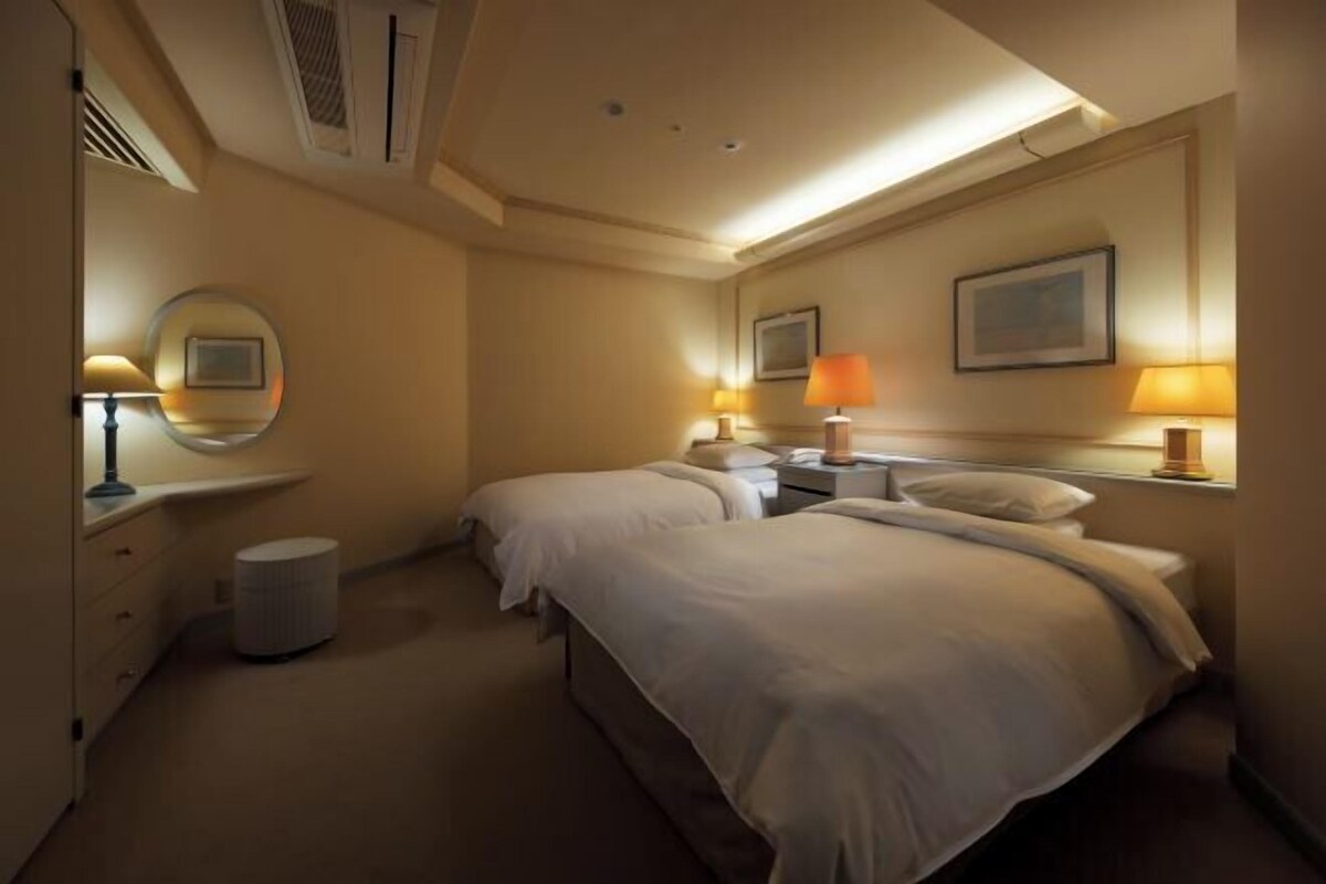 Habitación de hotel con dos camas, lámparas de noche, un espejo redondo, un cesto de basura pequeño y un cuadro en la pared.