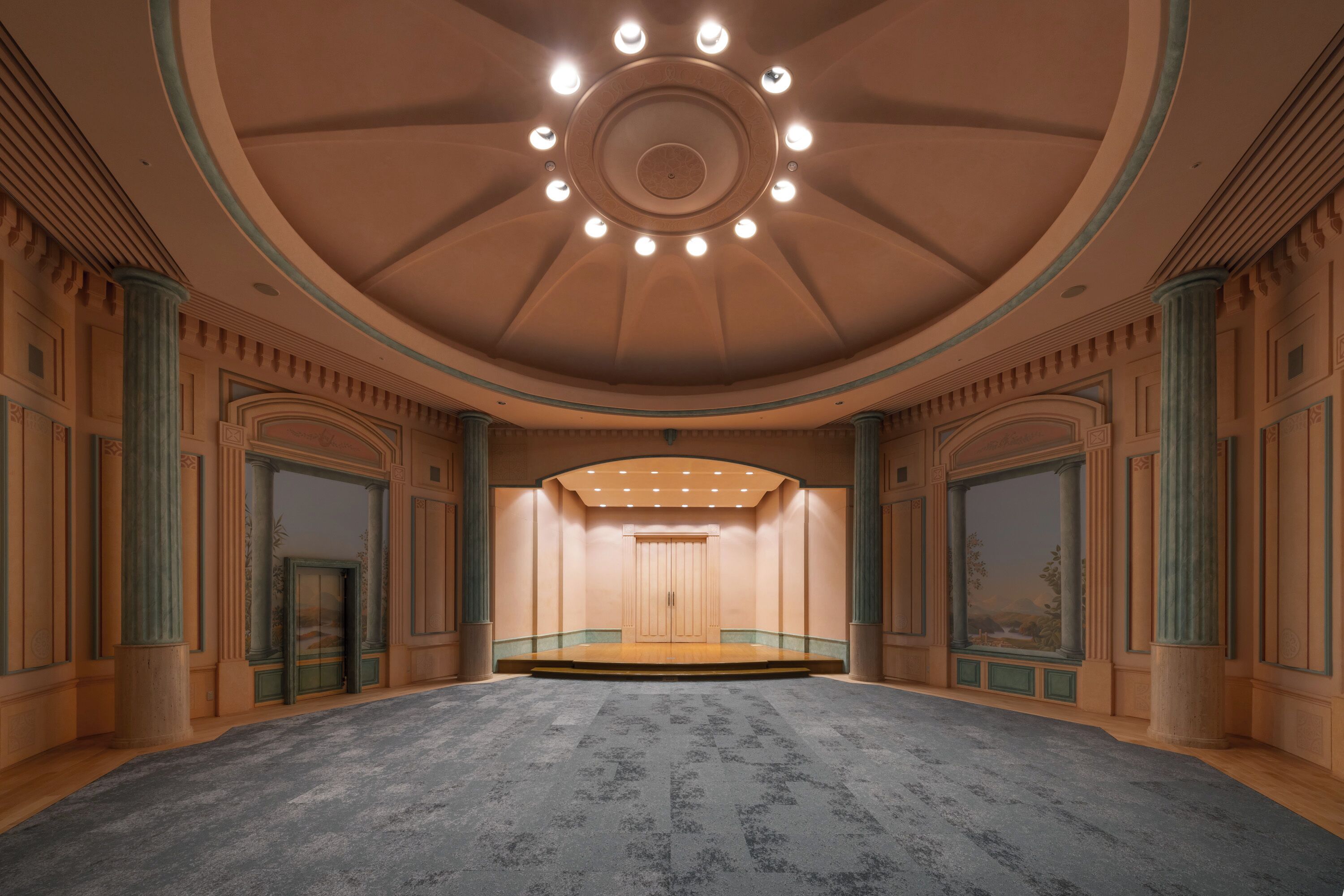 Une vaste salle dotée d’une scène centrale, de colonnes richement décorées et d’un plafond élevé éclairé par des luminaires disposés en cercle.