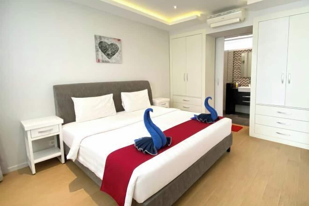 Une chambre d’hôtel avec un lit, des tables de chevet blanches, une armoire et une couverture de lit rouge.