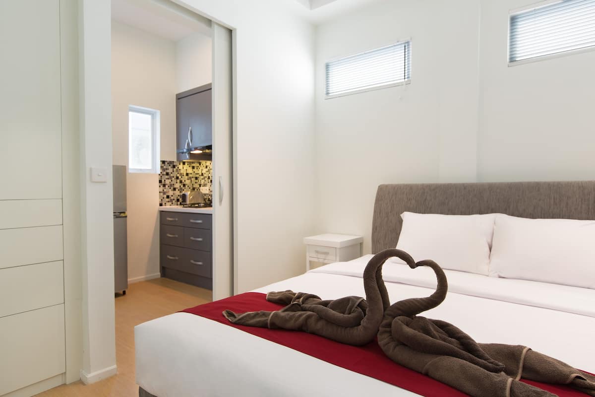 Une chambre à coucher avec un lit, une couverture rouge et une serviette marron disposées en forme de cœur. On y trouve une table de chevet blanche et une petite commode blanche placée à côté du lit.