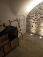 Una stanza con parete in pietra, termosifone bianco e scala di legno appoggiata al muro. Si nota un tavolo nero con una ciotola bianca sopra e scatole di legno etichettate con numeri.