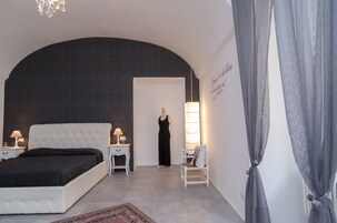 Una camera da letto con un letto bianco, un comodino con una lampada, una sedia e un manichino vestito di nero.