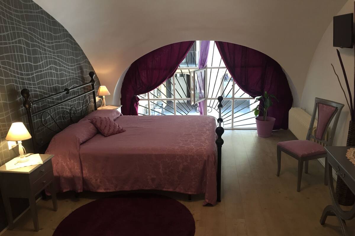 A bedroom with a curved ceiling, a bed with a headboard, a nightstand with a lamp, a chair, a desk, and a large window with curtains.