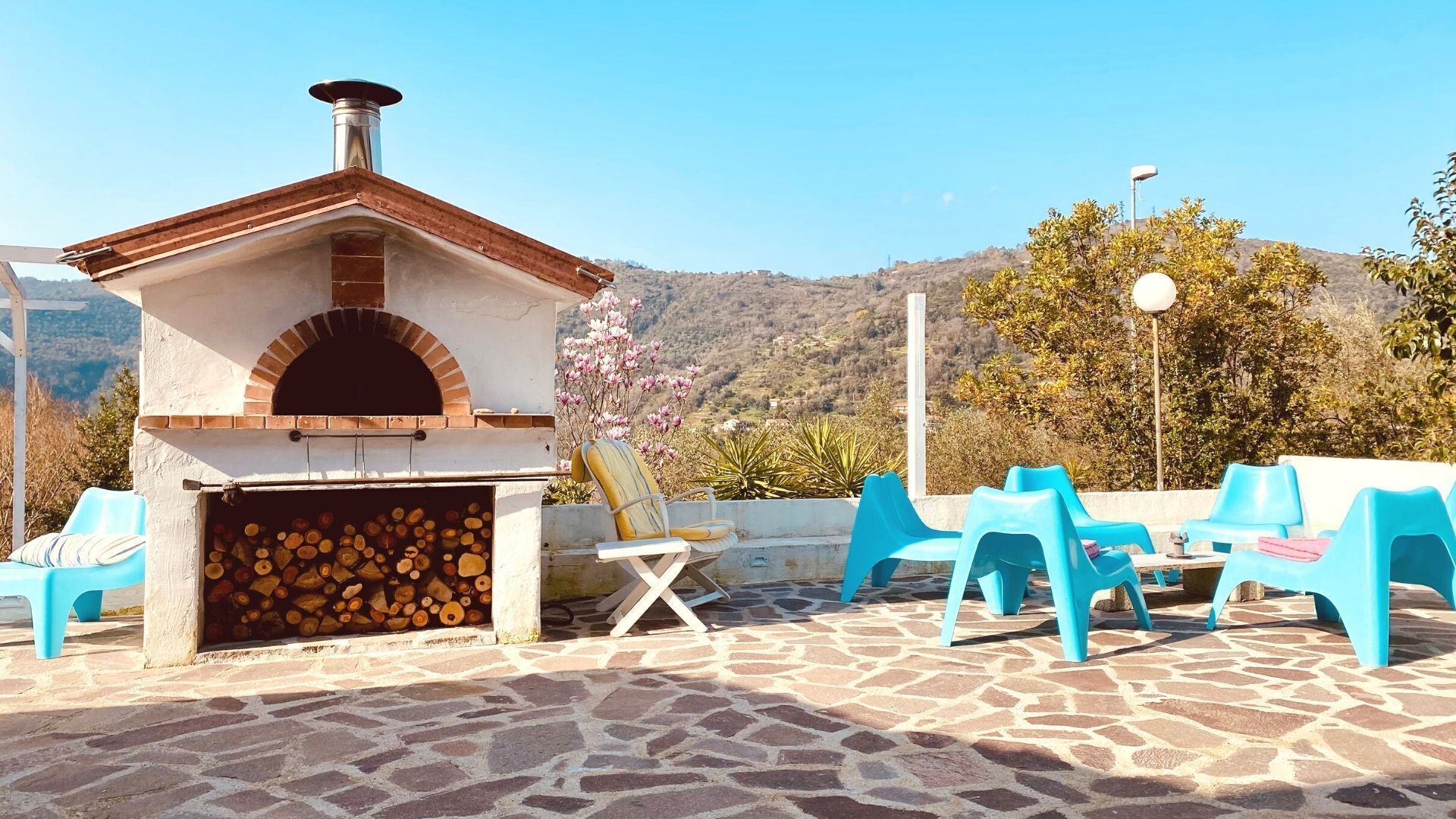 A stone patio with a brick oven, blue plastic chairs, and a view of hills.