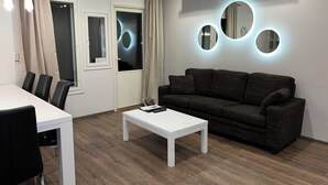 Una sala de estar moderna con una mesa blanca, sillas de cuero negro, un sofá marrón oscuro y una mesa de centro blanca. Hay tres ventanas con cortinas y las paredes tienen lámparas circulares.