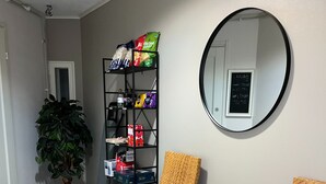 Una habitación con una silla de mimbre, una mesita con revistas, un estante con aperitivos, una planta en maceta y un espejo en la pared.