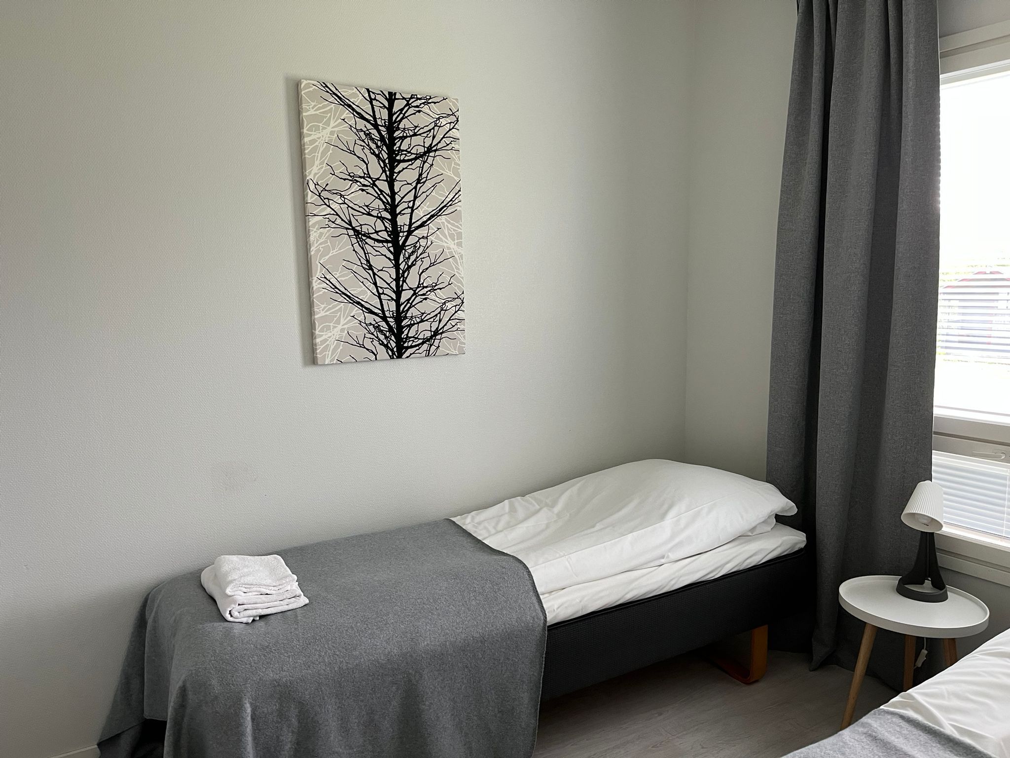 Pieni, siististi pedattu sänky, jossa on harmaa peitto ja valkoiset tyynyt, harmaa pyyhe lattialla, harmaa verho osittain vedettynä ja valkoinen pöytä, jonka päällä on lamppu.