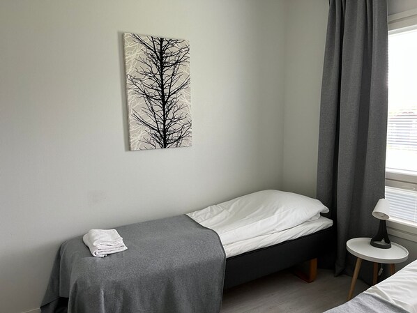 A small, neatly made bed with a gray blanket and white pillows, a gray towel on the floor, a gray curtain partially drawn, and a white table with a lamp. - Hotel Kontio (Kontiolahti)