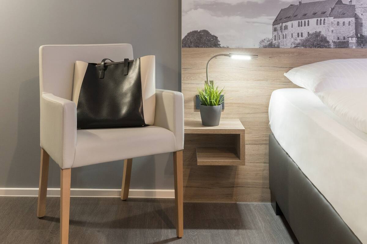 Un dormitorio moderno con cama, una silla con un bolso negro, una mesita de noche con una planta y un mural en la pared con un castillo.