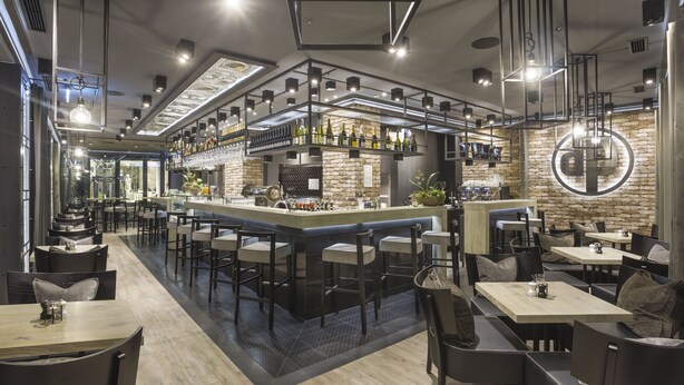 A modern bar area with a central counter, multiple seating options, and a stone accent wall. - dasPaul Hotel (Nuremberg)