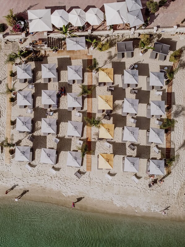 An aerial view of a beachside resort with multiple rows of umbrellas and lounge chairs, a sandy beach, and clear water.