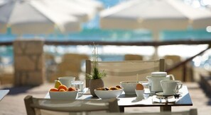 A table set with breakfast items, including fruits, a bowl of cereal, and a cup of coffee, with umbrellas and a view of the sea in the background.
