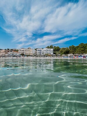 A coastal view with clear water reflecting buildings, a beach with umbrellas, and a row of multi-story buildings in the background.