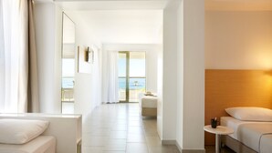 A hotel room with a bed, a sofa, a small table, and a view of the sea through a glass door.