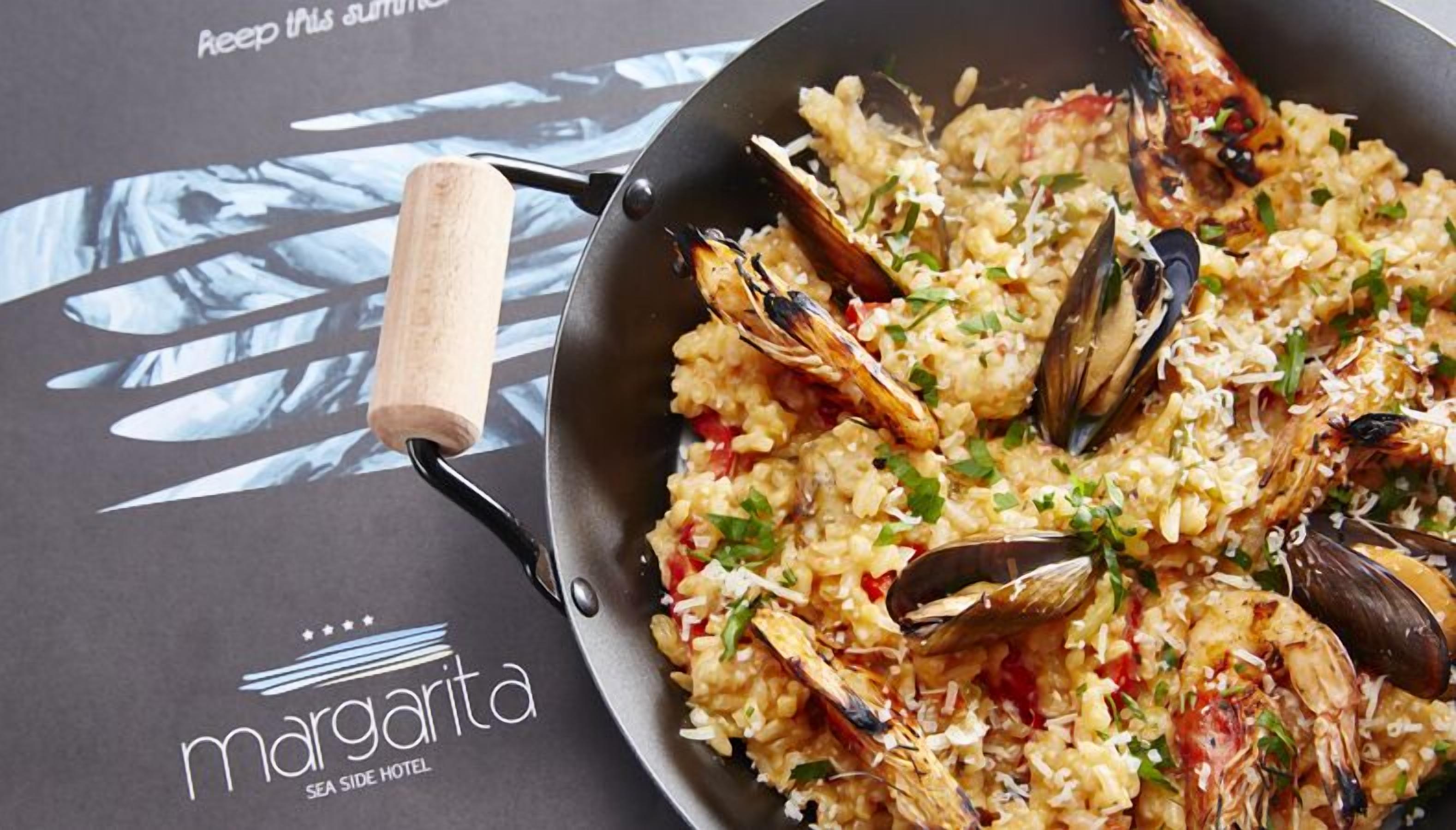 Una paellera de paella de mariscos con una sartén de mango de madera sobre una mesa con el logotipo y el texto que dice "margarita SEA SIDE HOTEL".