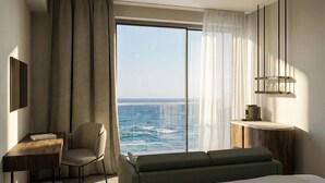 A modern hotel room with a large window offering a sea view, a comfortable sofa, a small table, and a minimalist decor style.