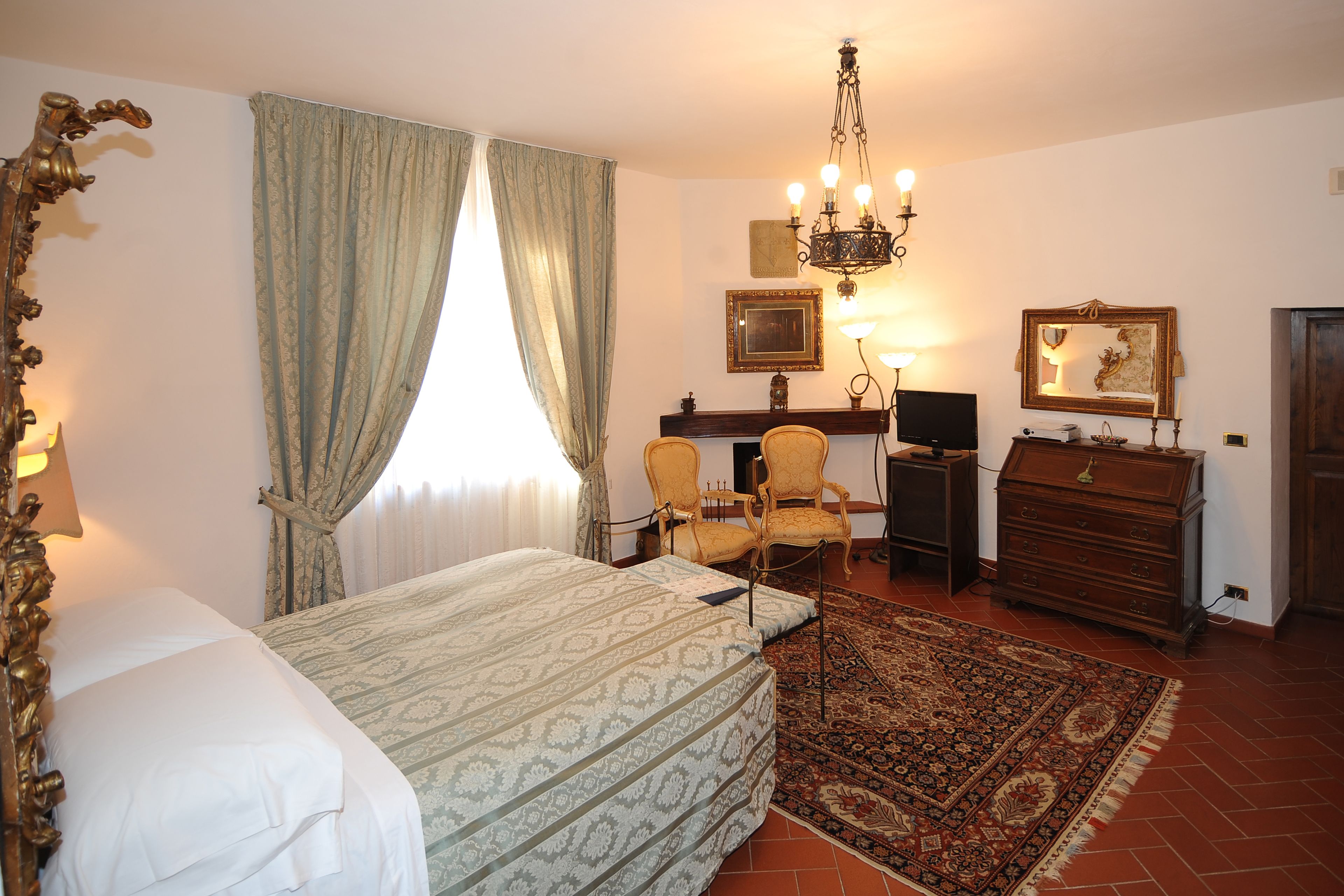 A hotel room with a bed, a desk with a chair, a dresser, a chandelier, and a large rug.