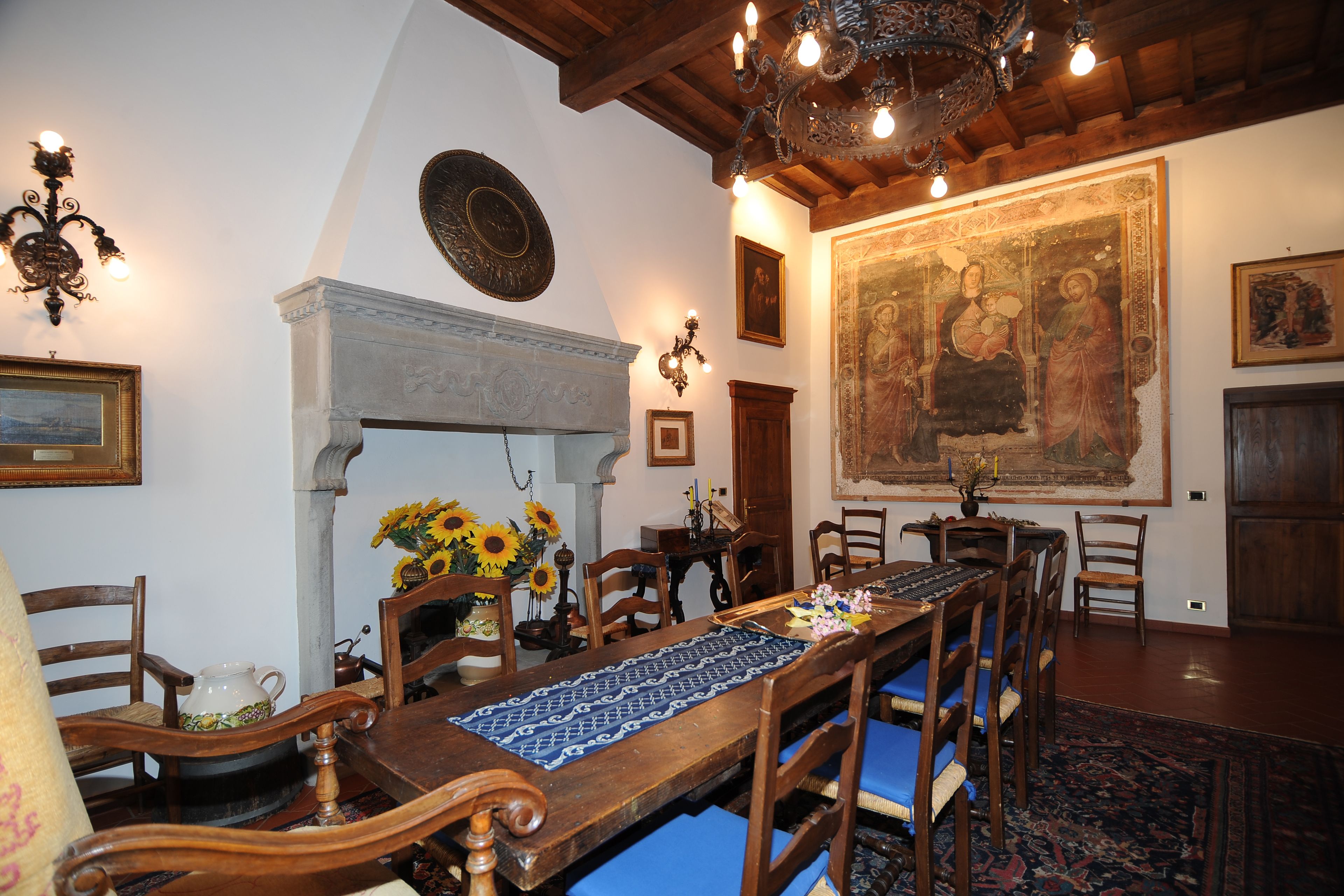 Une salle à manger avec une longue table en bois, des chaises rembourrées bleues, un lustre et un grand tableau religieux accroché au mur.
